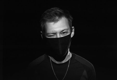 The guy in black clothes and a black medical mask on a black background winks and looks at the...
