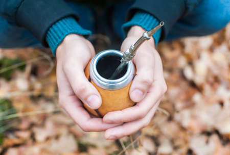 A man holds a traditional drink yerba mate in his hands. Outdoors in the autumn forest. Close-up. View from aboveの写真素材