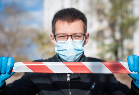 A man in a medical mask and gloves holds a signal tape in the street. Quarantine and isolation...