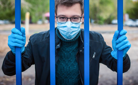A man in a medical mask and gloves behind a metal bars in the street. Quarantine and isolation...