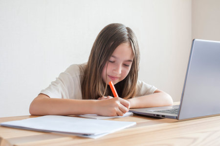 A little girl studies at a computer at home at a table with books and notebooks. Online training,...
