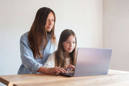 Young mother helping her daughter schoolgirl on distance learning session at home at table with laptopの写真素材