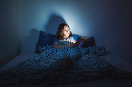 The girl lies at home in the evening on the bed with a phone in the dark. defocusの写真素材