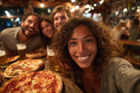 Happy woman with curly hair captures a selfie with friends at a vibrant restaurant, featuring pizzas and beer, showcasing a fun atmosphere of camaraderieの素材