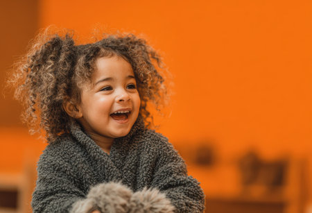 Happy young girl with curly hair, dressed in a soft gray sweater, is playing indoors against a bright orange backdrop, radiating joy and warmthの素材
