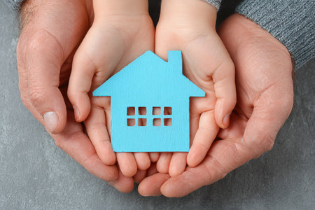 Adult and child hands cradle a blue wooden house symbol, embodying themes of family, love, and security in a warm, nurturing atmosphere with soft texturesの素材