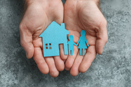 Hands are gently cradling blue paper cutouts of a house and family figures, representing warmth, security, and the essence of family life in a textured settingの素材