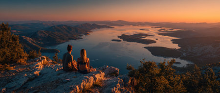 Two individuals enjoying a peaceful sunset view over a tranquil lake, with mountains and lush greenery creating a serene atmosphere and natural beautyの素材