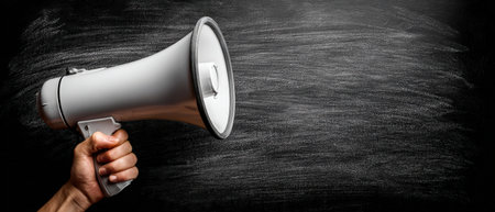 A hand grips a white megaphone in front of a textured black chalkboard, representing communication and the importance of voice in creative expression and announcementsの素材