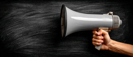 A hand grips a gray megaphone positioned against a textured black chalkboard, representing the essence of communication, announcements, and public speaking in an impactful wayの素材