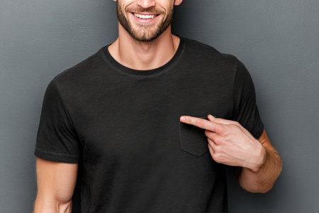 Confident man with beard, wearing black t-shirt, is pointing at his chest in a neutral background, creating an inviting atmosphere for personal branding and self-expressionの素材