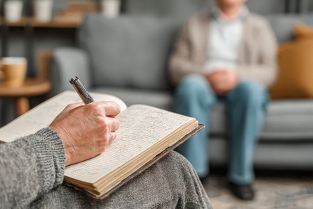 Senior man is sitting comfortably on a couch, while a therapist takes notes in a notebook, creating a supportive environment for discussionの素材
