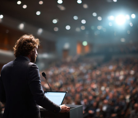 Male presenter is delivering a speech at a crowded conference, standing confidently at a podium, with attentive audience members in a modern auditoriumの素材
