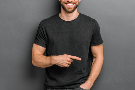 Male individual in a black t-shirt stands against a gray wall, smiling and pointing to the side, creating an engaging and inviting scene for marketing useの素材