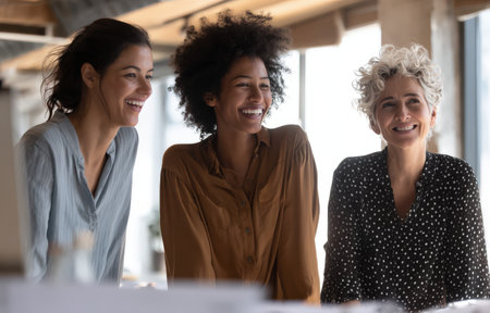 Three women with diverse backgrounds are smiling and interacting in a contemporary office setting, highlighting camaraderie and a positive atmosphere for teamworkの素材