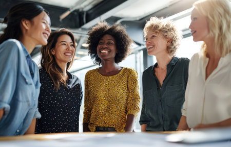 Diverse group of women smiling and interacting in a contemporary office space, highlighting friendship, teamwork, and positive energy in a professional settingの素材