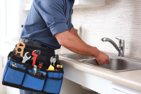 Male plumber in blue shirt is fixing kitchen sink, using tools from his belt, showcasing professional plumbing skills in a contemporary home settingの素材