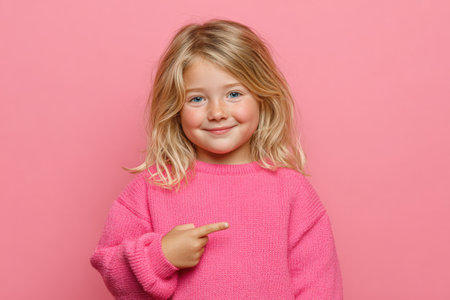 Cheerful young girl in a pink sweater smiles and points to the side against a bright pink backdrop, creating a playful and inviting sceneの素材