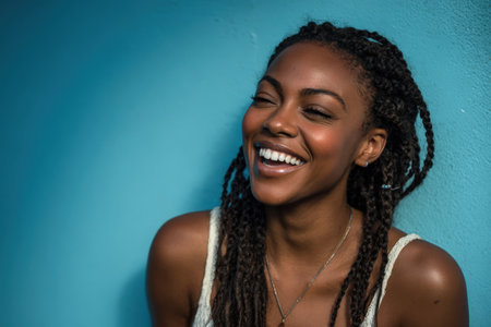 Joyful African American woman with braided hair is smiling brightly against a vivid blue wall, capturing a moment of happiness and warmth in a relaxed atmosphereの素材