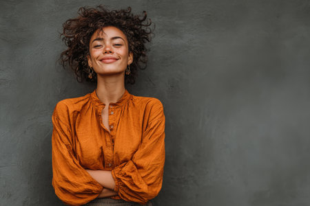 Confident young woman with curly hair in an orange blouse stands against a textured gray wall, radiating joy and positivity in a vibrant portraitの素材