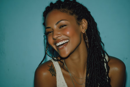 Joyful African American woman with dreadlocks, wearing hoop earrings, smiles brightly against a blue backdrop, showcasing her vibrant personality and carefree spiritの素材