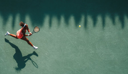 Female tennis player is leaping on green court to hit a ball, demonstrating skill and agility, with shadows creating a dramatic effect in the backgroundの素材