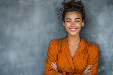 Confident woman with curly hair in an orange blouse stands against a textured gray wall, radiating warmth and positivity in a casual yet engaging mannerの素材