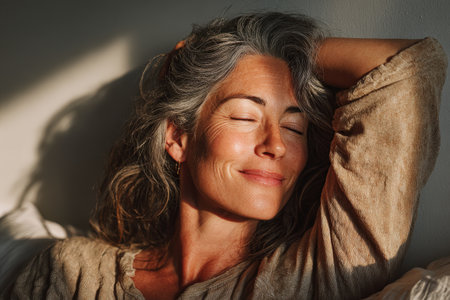 Woman with gray hair is relaxing on a sofa, basking in warm sunlight, creating a serene atmosphere filled with comfort and tranquility in a cozy indoor settingの素材