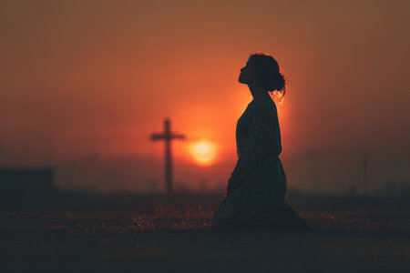 Woman in silhouette kneeling in prayer at sunset, with a cross behind her, creating a peaceful and reflective ambiance, evoking themes of spirituality and devotionの素材