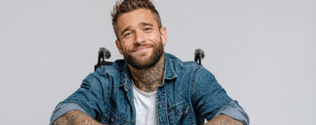 Confident man with tattoos, wearing denim jacket, seated in wheelchair, radiating positivity and strength in a contemporary studio environment with neutral backdropの素材