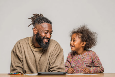 Smiling African American man and girl share a joyful moment at a table with a tablet, highlighting their connection and engagement in a cozy environmentの素材