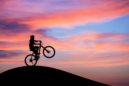 silhouetted mountainbiker doing wheelie in sunset sky on hillの写真素材