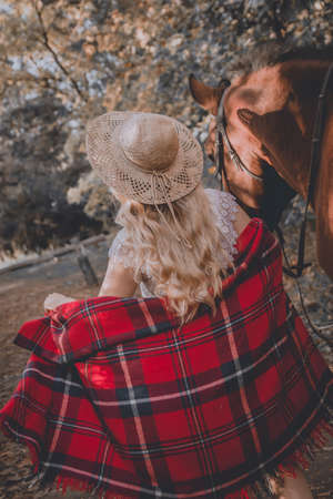 Blonde girl in hat and plaid walks with horse in the evening on a farm in the villageの写真素材