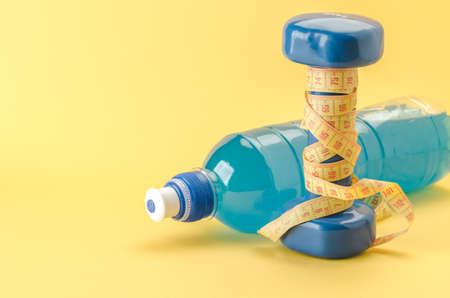 Fitness concept with dumbbell with yellow centimeter and bottle/blue dumbbell with centimeter and bottle on a yellow background. selective focus. Copy spaceの写真素材