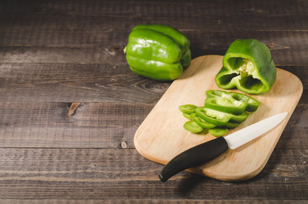 Sliced green paprika pepper on a dark wooden table/Slicing peppers on a wooden cutting board with a white knife with the black handle, copy spaceの写真素材