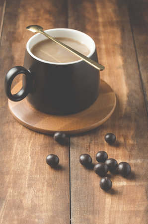 black mug of a cappuccino with a spoon in a wooden support with the chocolate drops scattered on a wooden table.の写真素材