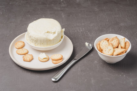 Cheese and cracker in a white bowl with a knife on a dark stone background.