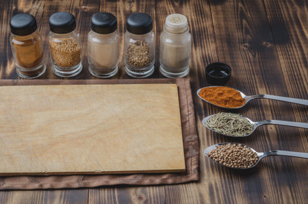 Various spices in spoons, round cutting board and cooking utensils on wooden background.