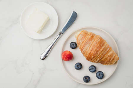 Croissant with berries in white bowl and butter knife on white stone background. Top viewの写真素材