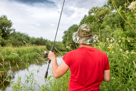 Fisherman in a panama hat and a red t-shirt casts a spinning rod. view from the back. hobby fishing.の写真素材