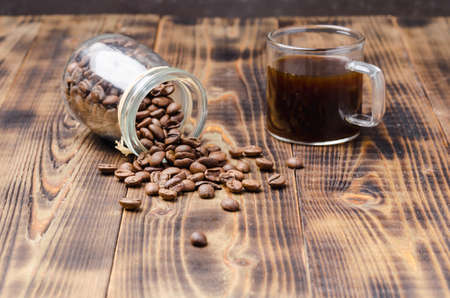 Cup of coffee and beans are scattered from a glass jar on a wooden table.の写真素材