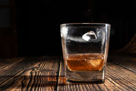 Whiskey with ice cubes on a wooden table. An old tabletop with light and a glass of strong drink. A splash of whiskey in a glass while serving ice.の写真素材