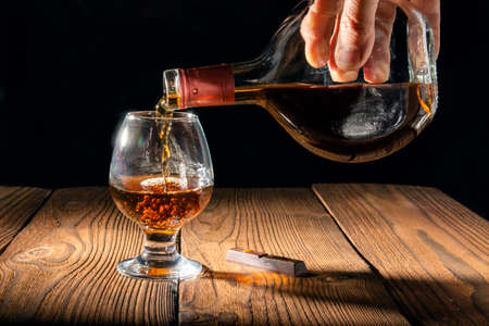 Pour cognac or brandy into a large glass on an old rustic table. Vintage wooden background, night mood. Place for text, dark background, selective focus.
の写真素材