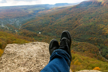 Male legs in sneakers sit on the edge of a high cliff and watch the mountains and forest on a sunny autumn dayの写真素材