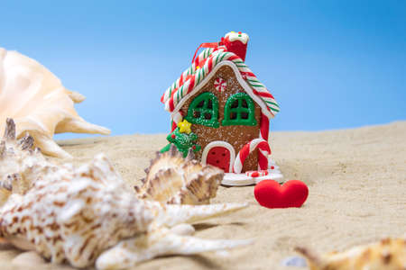 A small Christmas toy in the shape of a gingerbread house stands on a sandy beach next to beautiful shells. Merry Christmas by the warm sea.の写真素材