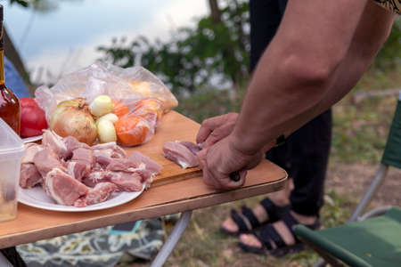 Dinner al fresco by the river. Portable folding table and picnic chairs. A man cuts meat to prepare a roast.の写真素材