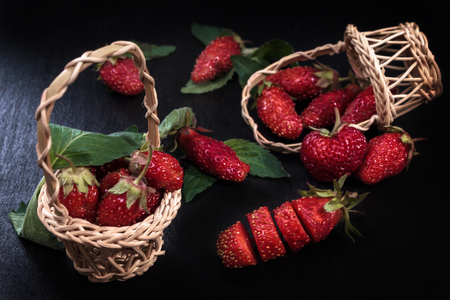 Mature red strawberries on a black table and fresh ripe strawberries in a wooden basket. Red on blackの写真素材
