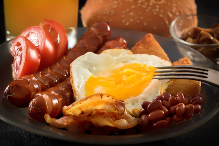 Full English breakfast with scrambled eggs, bacon, sausage, beans, tomatoes and juice. fork pierces the egg yolk. Black backgroundの写真素材
