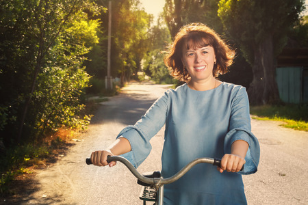 Joyful young woman walks next to a bicycle on a country road in the countryside in the rays of the sunset. Tonedの写真素材