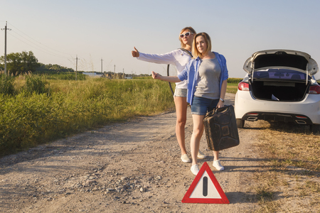 Two driver girls vote on a rural road in anticipation of help with an empty fuel tank and a hose near the car in the rays of the sunset. Toned. Copy spaceの写真素材